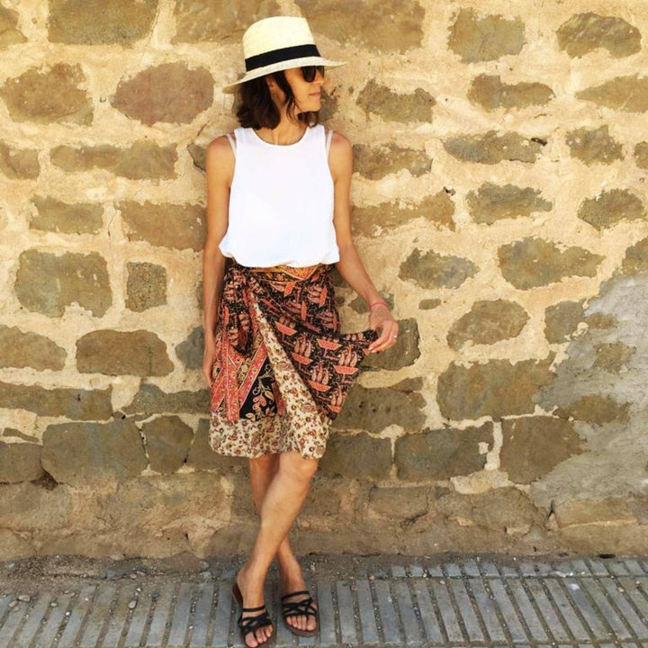 A woman standing in front of a stone wall in Italy. She's wearing a Black Sari Wrap Skirt with Tan and burnt red accents. She's paired the skirt with a white tank top and strappy black sandals.