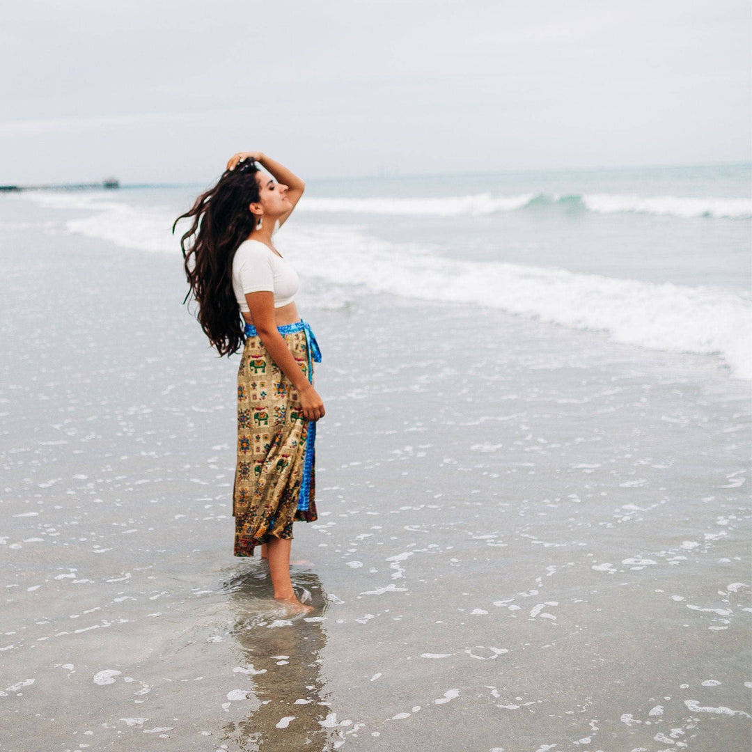 A woman standing with her feet in the ocean feeling the breeze on her skirt. She's holding up her Sari Wrap Skirt away from the water. Her skirt is tan with little blue elephants and tribal patterns. The edges are light blue. She's paired it with a white crop top and chunky gold jewelry