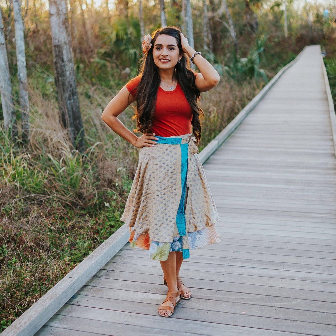 The model is standing on a board walk on a warm summer night. She's wearing a tan colored Sari Wrap Skirt with Light blue accents. The skirt has a peacock feather pattern on it. She's paired the skirt with a red tee and tan sandals.