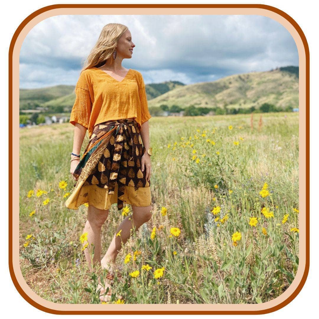 Model stands in a field of flowers. She wears an orange top and a sari wrap skirt. The skirt is dark brown with a light tan and brown pattern that flows throughout the skirt.
