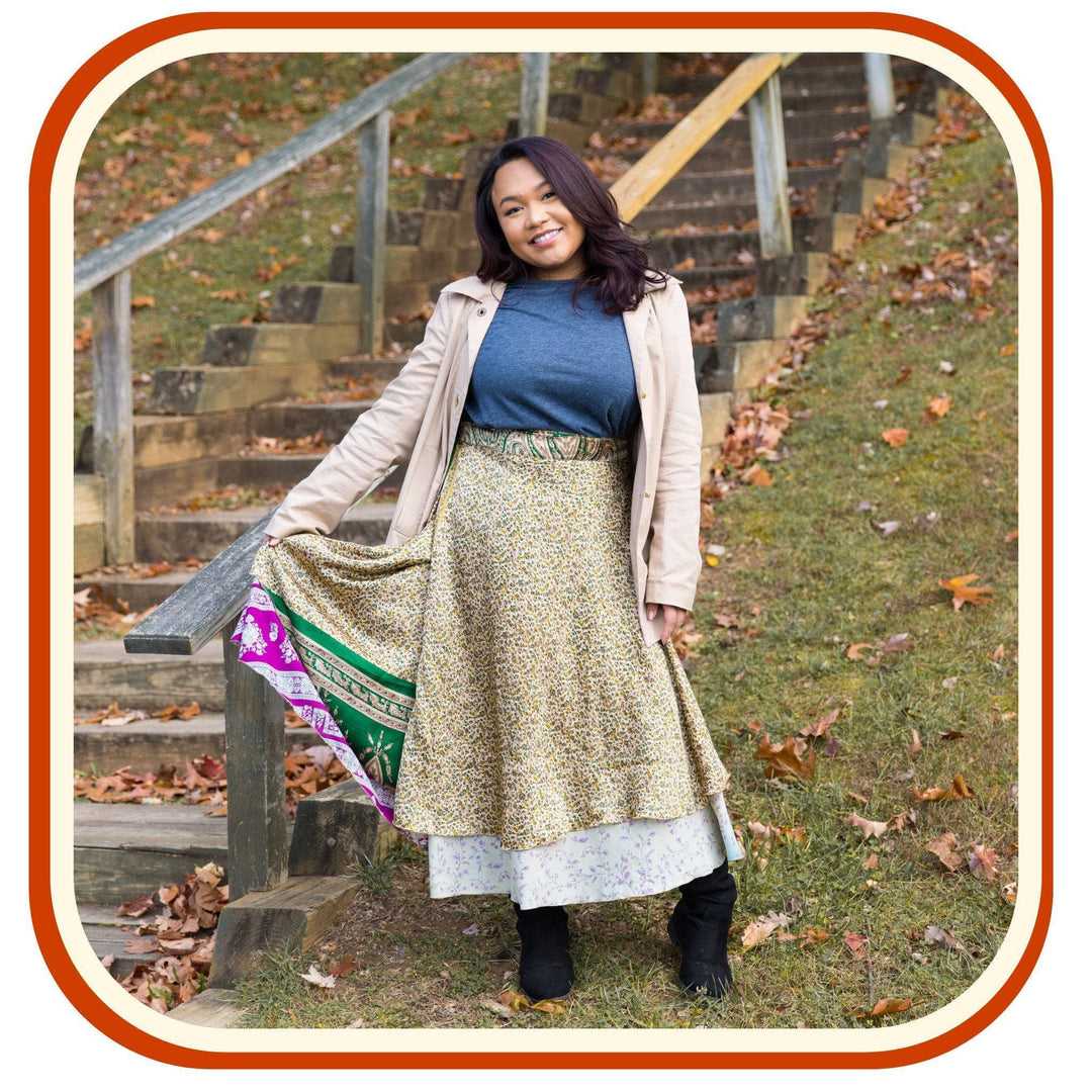 Model standing below a staircase with leaves all around her. She is wearing a Sari wrap skirt that is a dusty green and a light sky blue with a green and pink pattern underneath the skirt. She also wears a pale pink jacket, leggings and a blue t shirt.