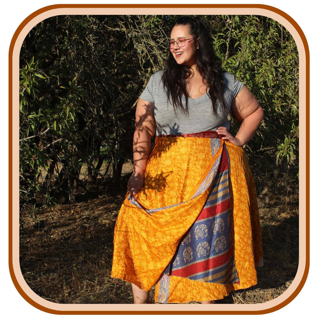 Model stands against trees. She wears a light grey t shirt and a bright light orange sari wrap skirt. The skirt has a subtle light orange texture on it. The second layer of the skirt is a dark blue, red , tan, and grey pattern.
