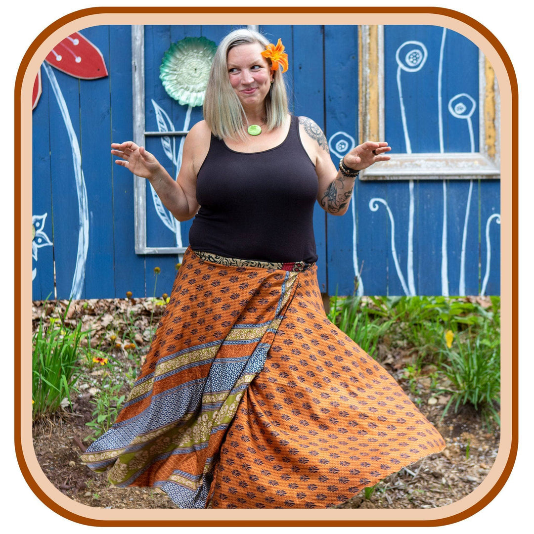 Model stands against a blue mural outside. She wears a black tank top, an orange flower in her hair, and an orange sari skirt. The skirt is orange with a navy blue pattern on it. The skirt also has a white and blue section towards the bottom of the skirt on the second layer.