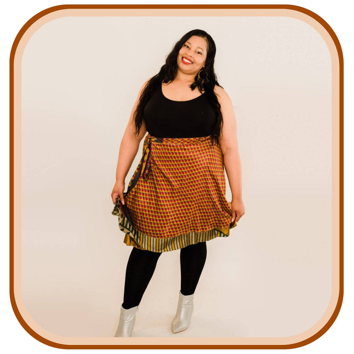 Model stands against a white background. She wears a black top, silver shoes, black leggings and an orange sari skirt. The sari skirt is orange with a red pattern on it. The skirt also has a yellow trim with black stripes on it.