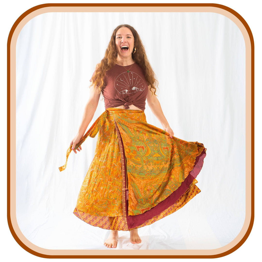 Model stands against a white background. She wears a light brown t shirt, and a sari wrap skirt. The skirt is a light orange with green and dark orange patterns on it. The skirt also has a maroon layer underneath of the light orange layer.