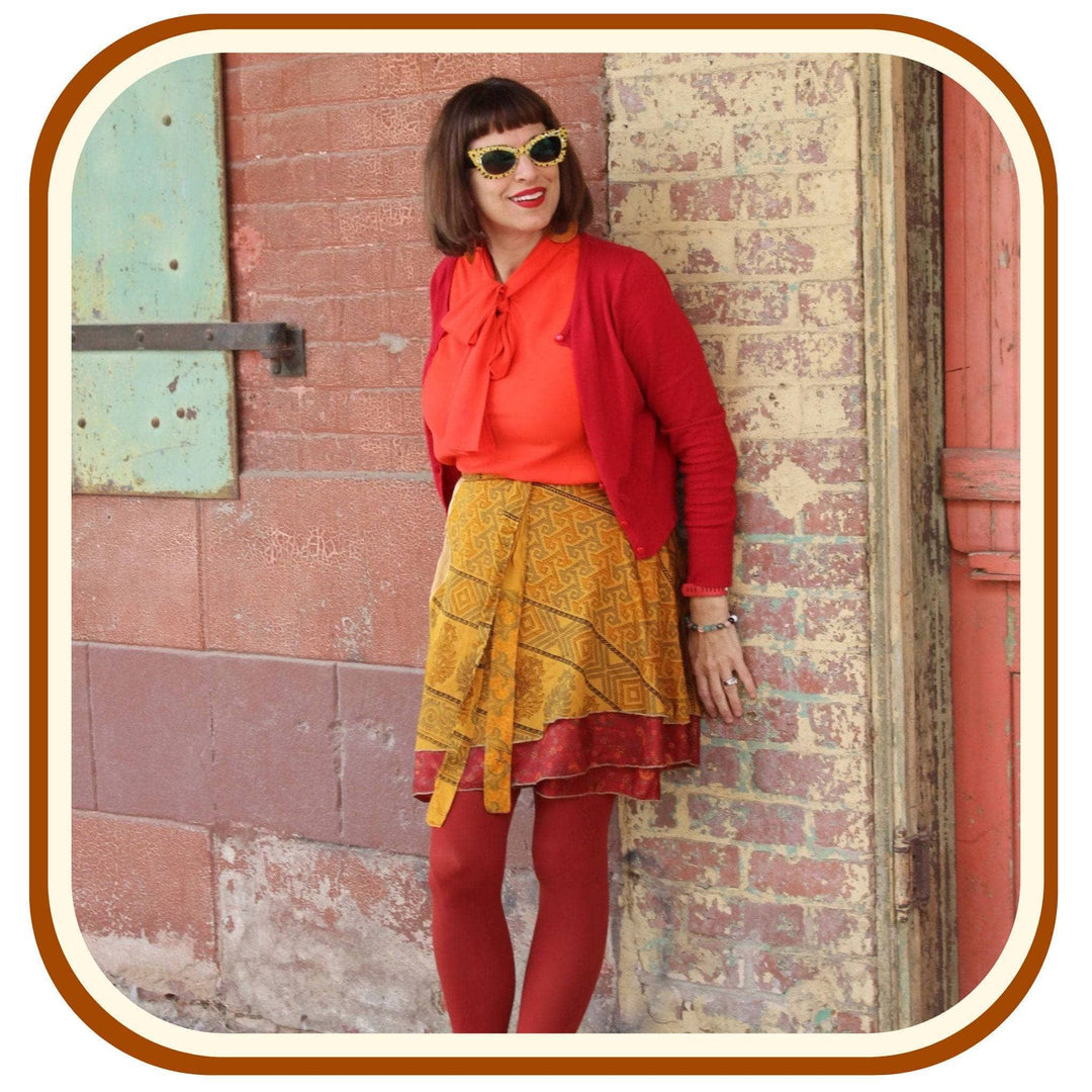 Model leans against a brick wall. She wears a bright orange top with a red jacket. She also wears red leggings and a sari wrap skirt. The skirt is a dusty yellow with a red trim at the bottom. The pattern on the skirt is made up of brown, and tan.