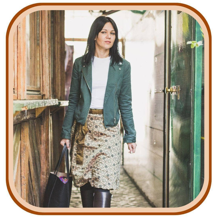 Model stands in a rustic doorway. She wears a green jacket with a white top, black boots, a black hand bag, and a sari wrap skirt. The skirt is a light tan with a black pattern that looks checkered with a minimal pattern throughout. The skirt has a gold belt.