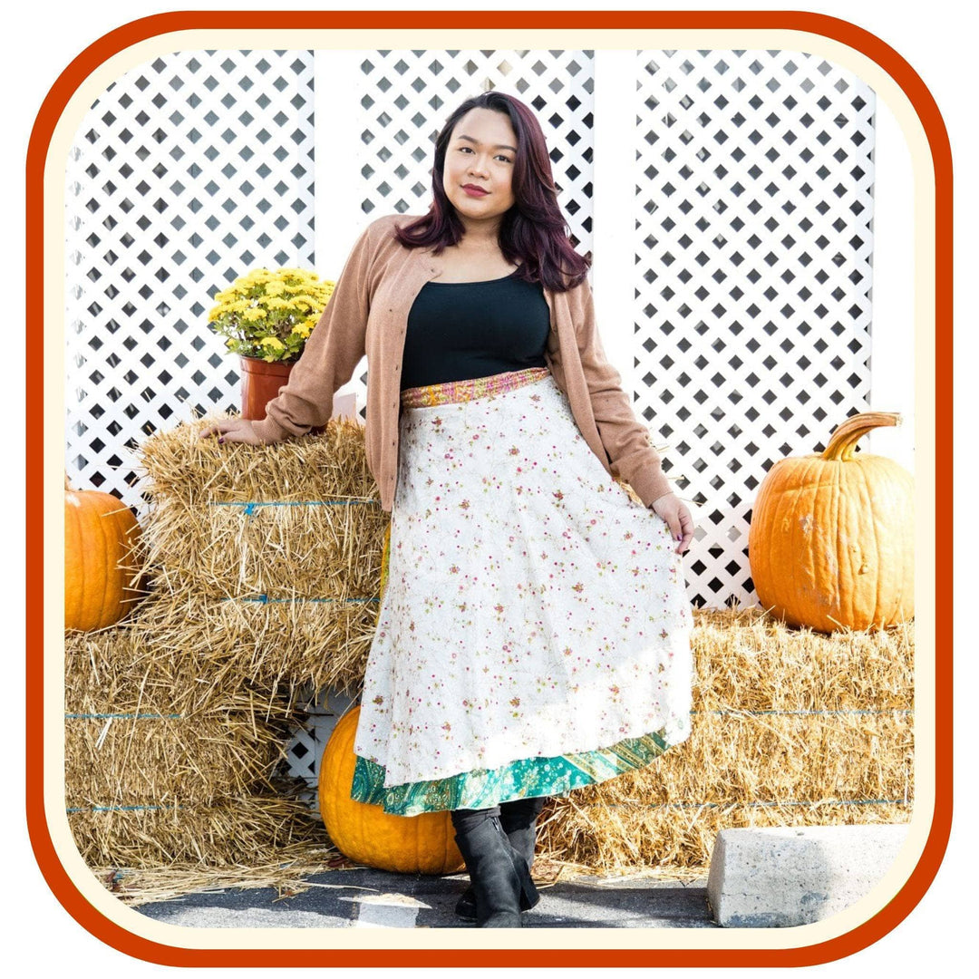 Model leans against hay bails with pumpkins on them. She wears a light brown jacket, a black top, black boots, and a sari wrap skirt. The skirt is white with a dark orange floral pattern that is very minimal. The skirt has a green trim on the bottom and an orange trim on the top of the skirt.