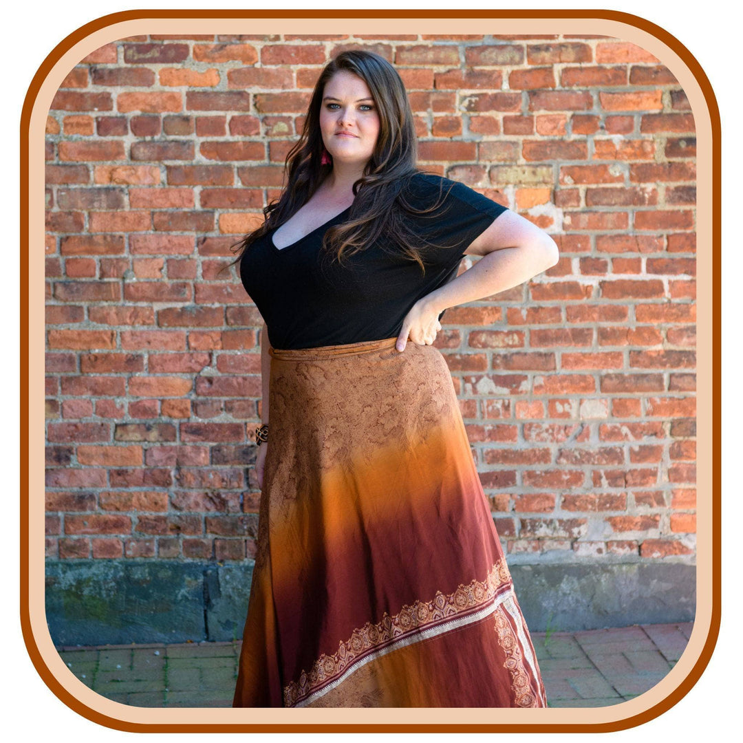 Model stands against a brick wall. She wears a black top and a sari wrap skirt. The skirt is a light tan, orange-brown and a dark brown. There is a small pattern on the skirt in white.
