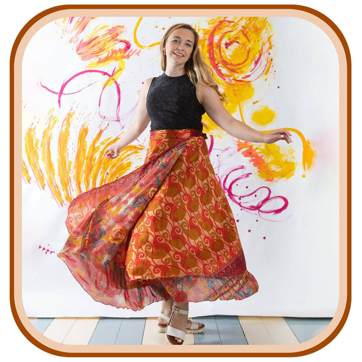 Model stands against a colorful painted background. She wears a black tank top, white sandals, and a sari wrap skirt. The skirt is a light orange with a dark orange pattern on it.