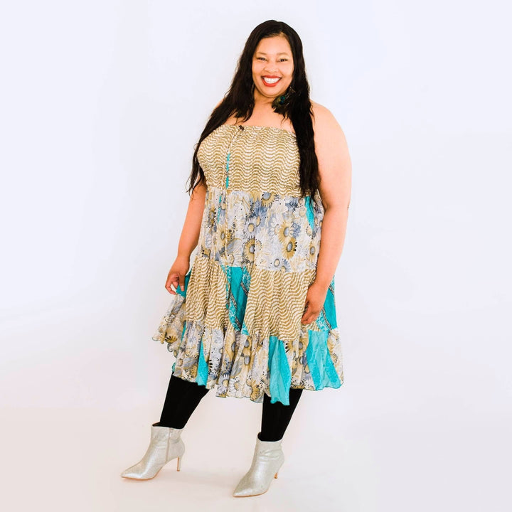 A woman standing in front of a white backdrop wearing a cream and baby blue plus sized sari patchwork dress. She's wearing it with leggings and a silver boot. The patchwork dress has floral patterns all over it.