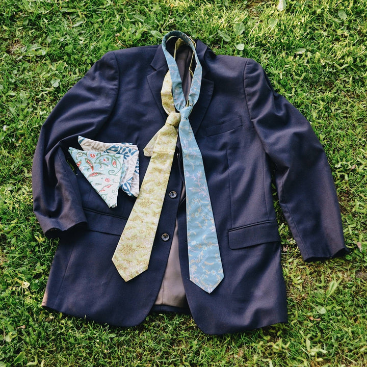Two silk neck ties and two pocket squares laid on top of a suit jacket on grass. 