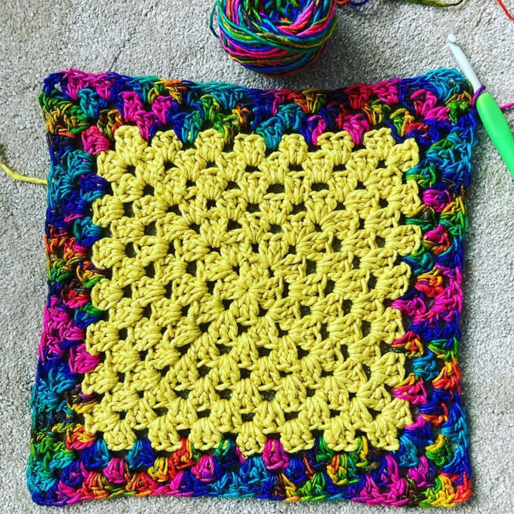 Crochet granny square made from silk roving worsted weight in illuminating and watercolors in front of a light grey background. Creen crochet hook and the rest of the watercolors skein are also visible.