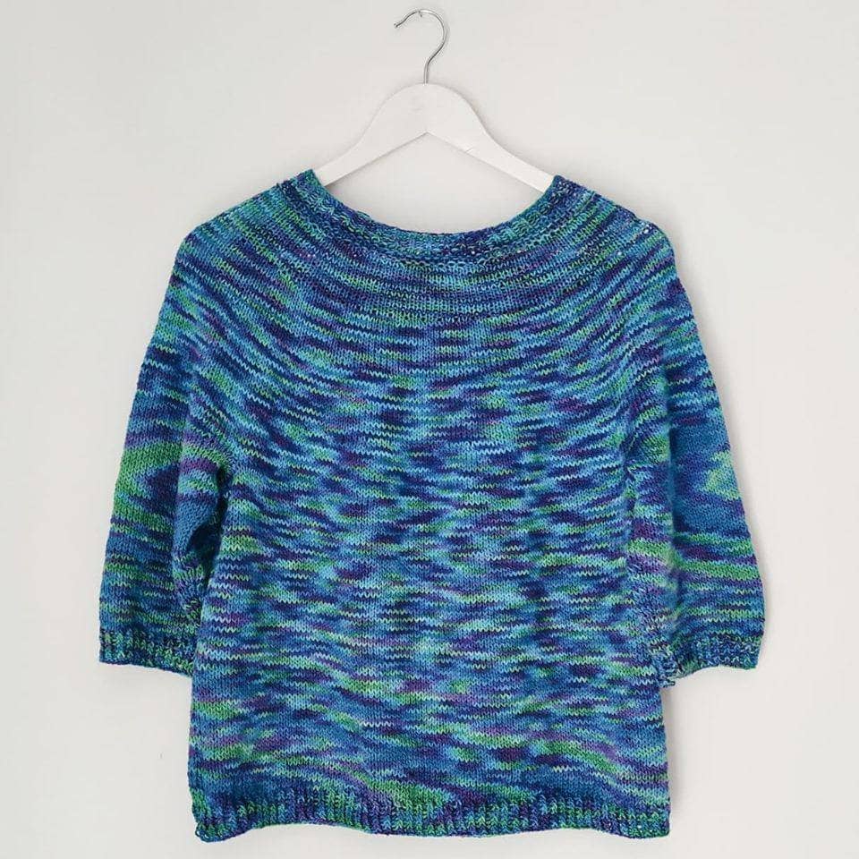 Simple seamless sweater in colorway peacock (blue, green, and purple) on a hanger in front of a white background.