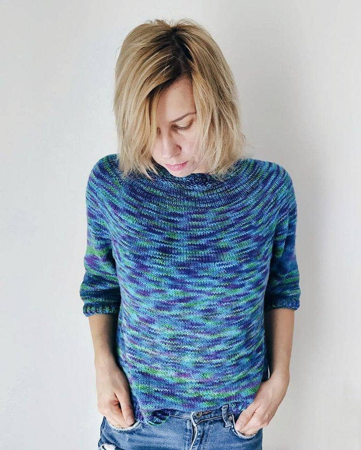 Designer wearing simple seamless sweater in colorway peacock with jeans in front of a white background.