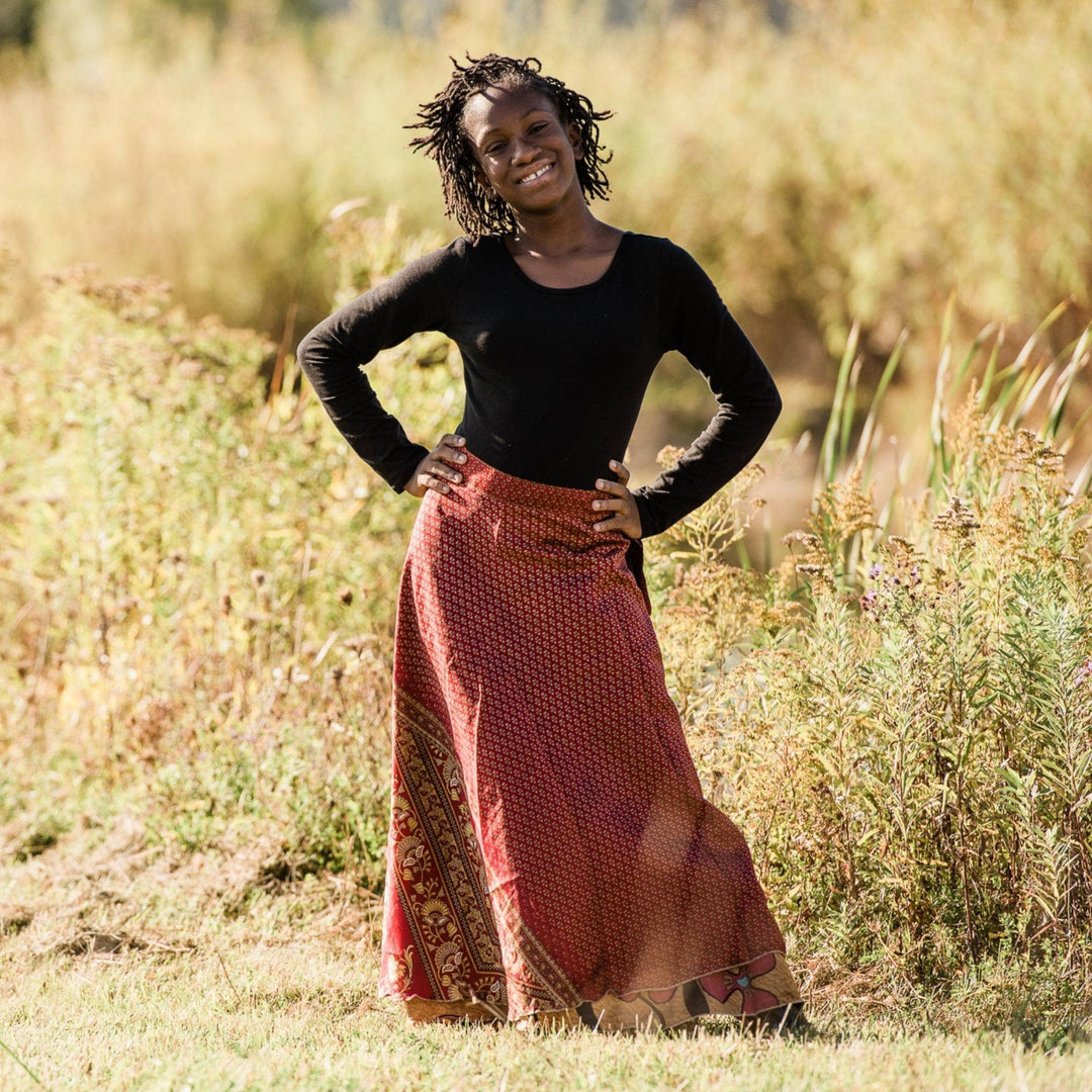 A junior model is wearing a red maxi sari wrap skirt while standing in front of tall grass. 