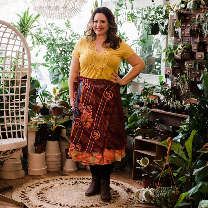Model is wearing a maroon and orange sari wrap skirt with potted plants in the background.  