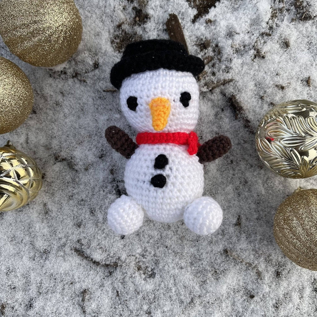 Snowman Amigurumi  with a red scarf and a black top hat laying in the snow with Christmas ornaments around it. 