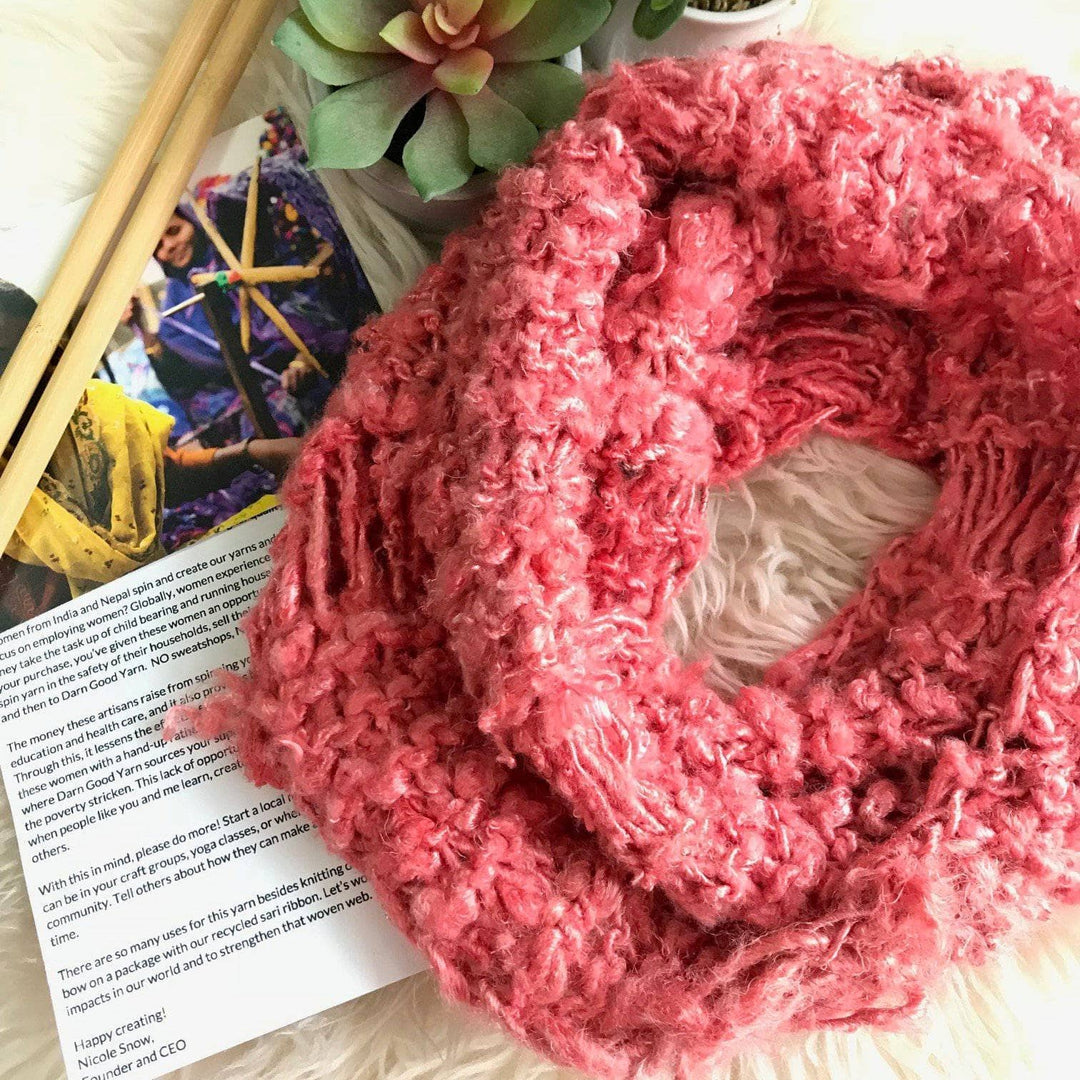 Spring Infinity Scarf in Coral sitting on a white fur background with knit pattern, knitting needles, and a small plant