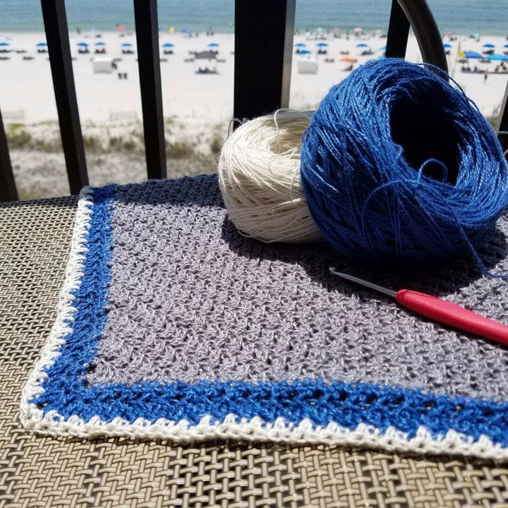 Low Tide shawl being created with crochet hook and two skeins of yarn (white and blue) with a beach in the background.