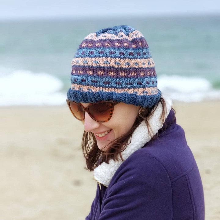 Woman outside wearing a blue, purple and pink matching striped glove and hat set on a beach