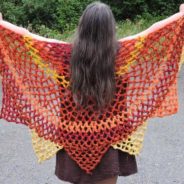 Woman holding the Sunburst Shawl in red and orange behind her back in front of greenery