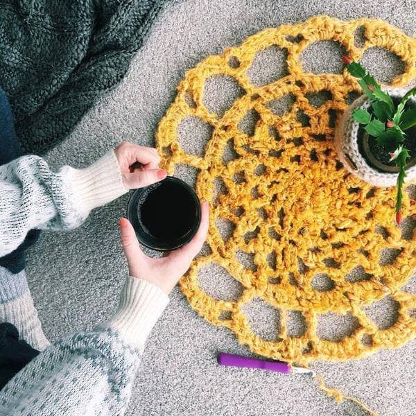Sunshine Mat in Sunflower (yellow) on a gray surface with a houseplant, woman's hands holding coffee, and a crochet hook