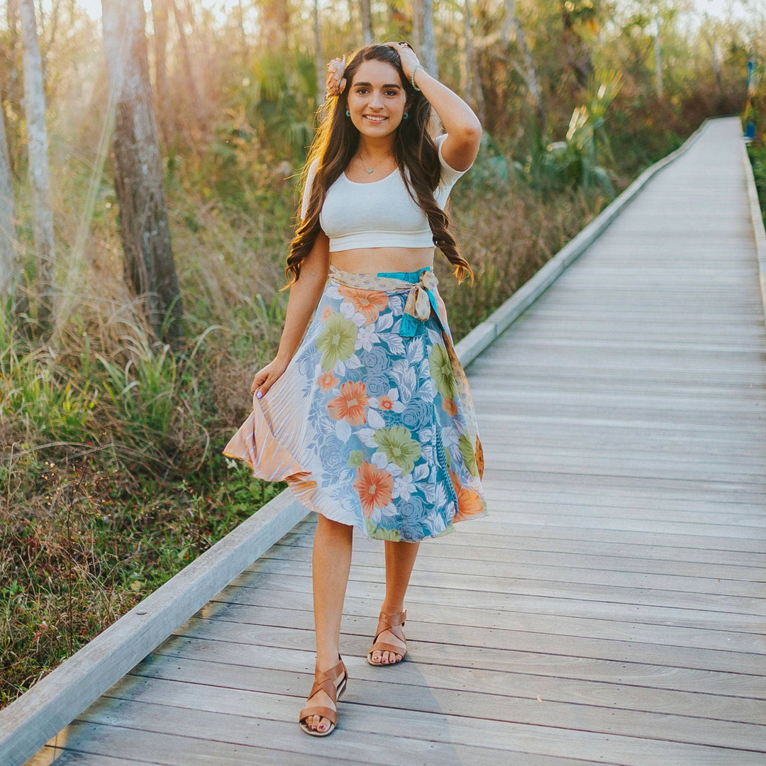 A girl standing on a boardwalk wearing a 00-04 sari wrap skirt in a tea length. It's a neutral color with blue and orange flowers all over it. She's wearing a cropped white tee and a flower in her hair.