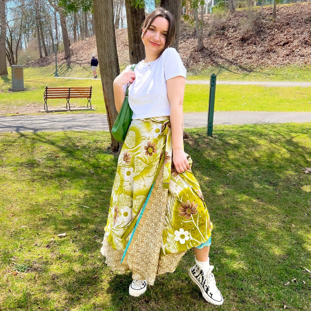 A woman standing in a park wearing a petit sized maxi length wrap skirt made from reclaimed sari. The skirt is light green with brown and white flowers.