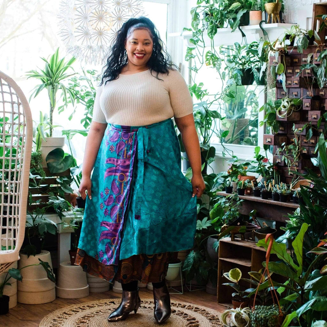 A woman standing in a floral shop. She's wearing a Goddess sari wrap skirt in a tea long. The skirt is teal with lavender accents.. She's wearing a tall leather boot and a cream colored tee with it.