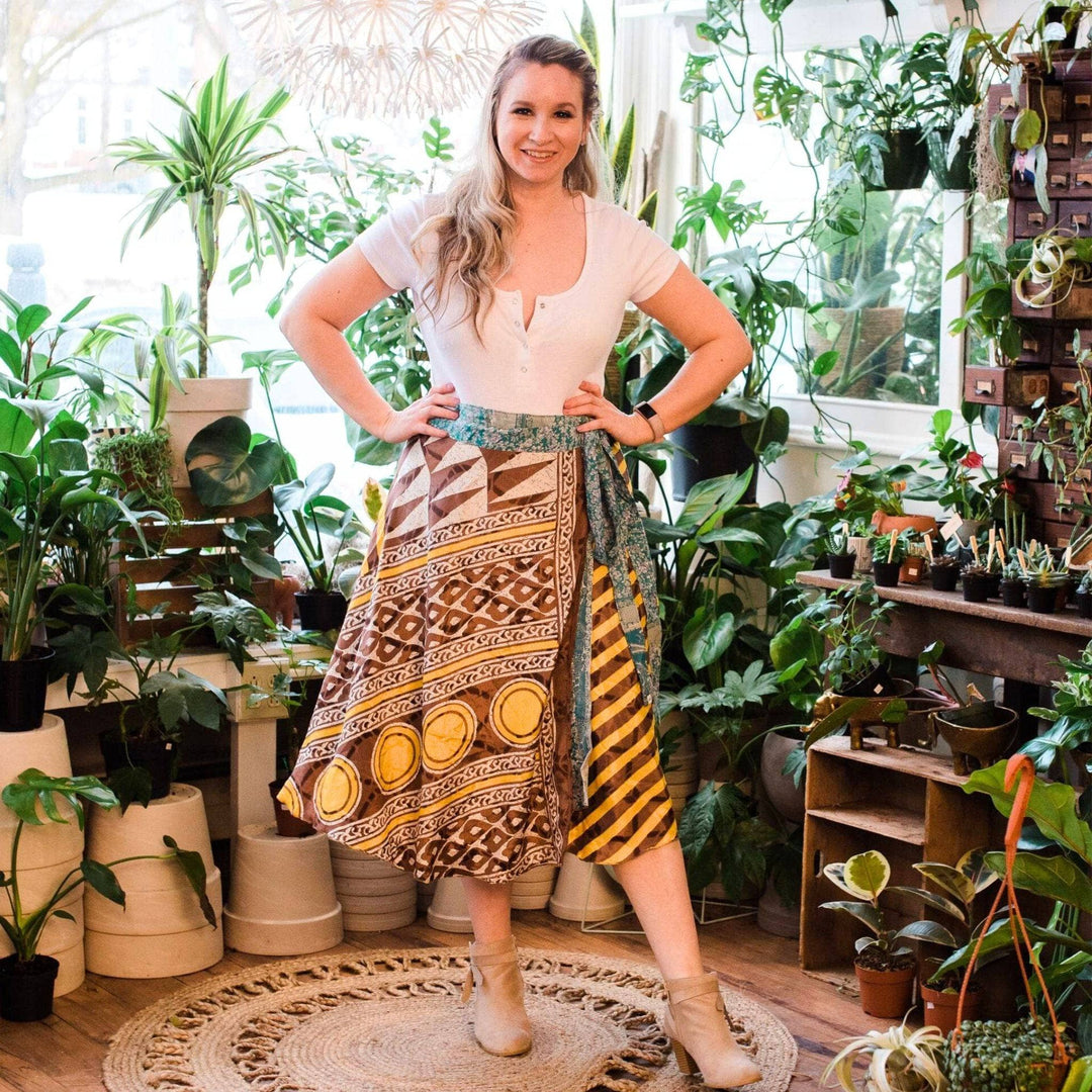 A woman in a flower shop wearing a tea length reclaimed sari wrap skirt. The skirt is cream, brown and gold shapes with green accents. She's wearing a white colored top with it.