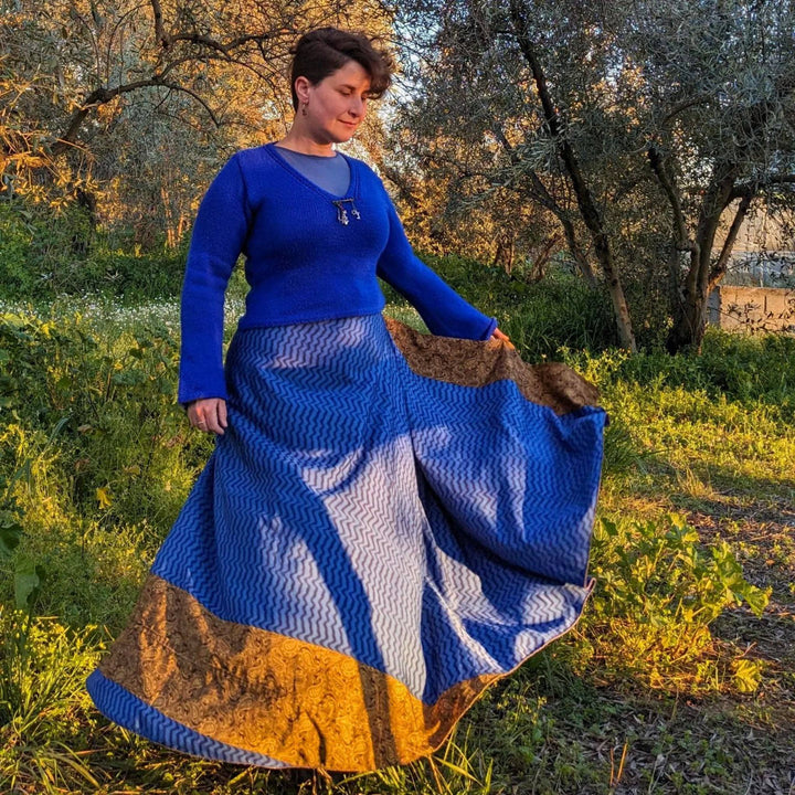 A woman standing in a field wearing a saphire reclaimed sari wrap skirt. She's wearing a 12-18 sized sari wrap skirt in a maxi length. The skirt is blue with a gold strip along the bottom, she's paired in with a matching sweater.