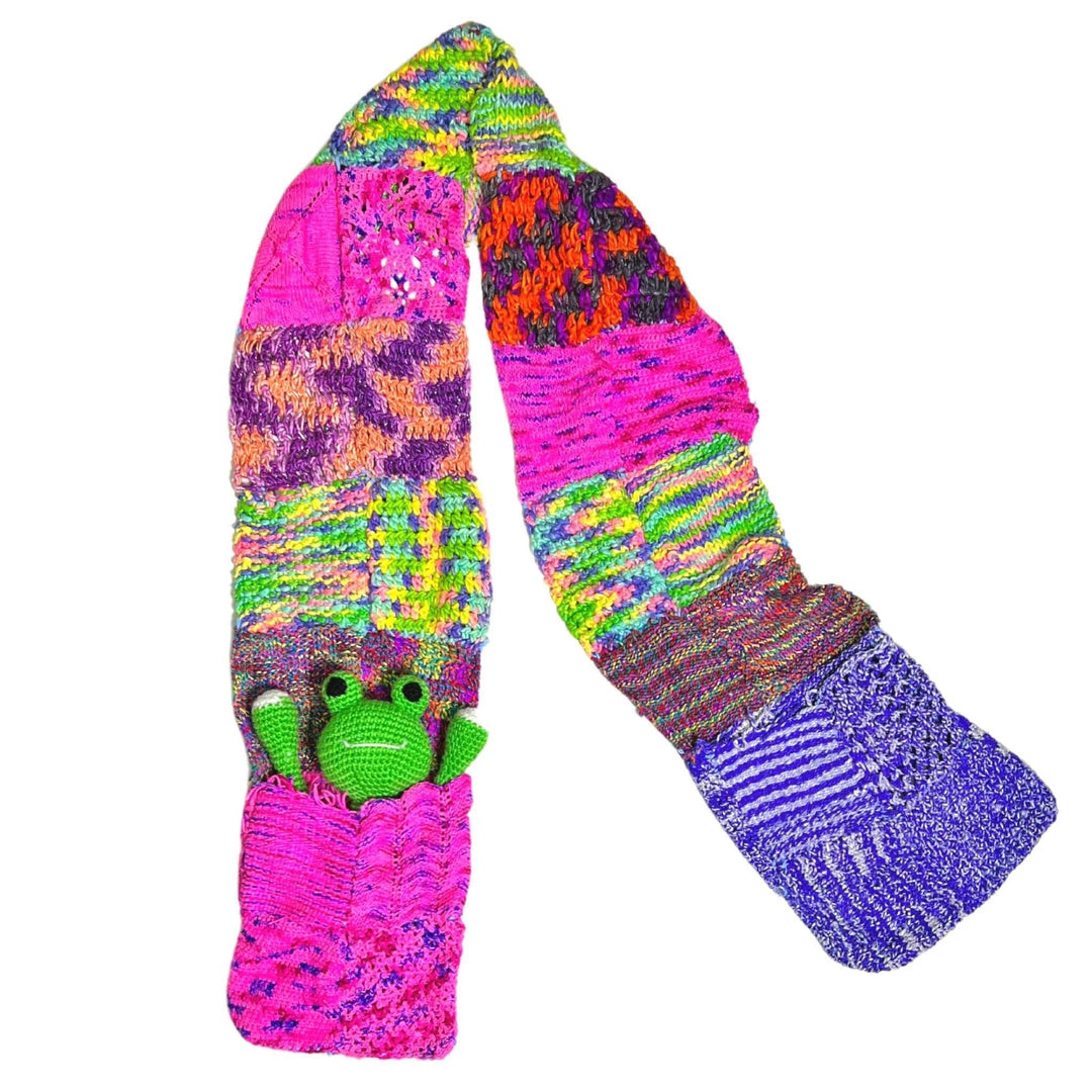 Patchwork scarf (multicolor) with pockets and amigurumi frog in one of the pockets laying on a white background.
