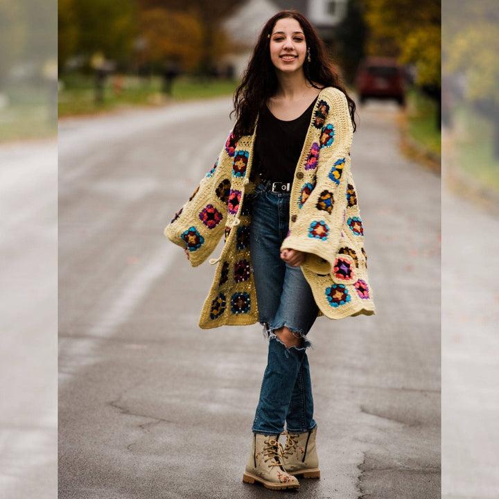 Model wearing regular size technicolor long cardigan (crochet granny square, beige and multicolor) while standing outside on a quiet street.