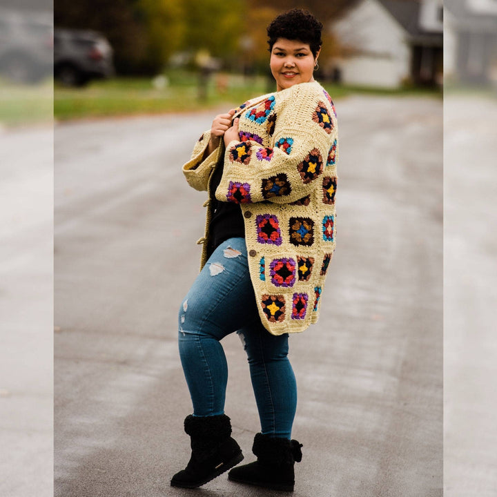 Model wearing goddess size technicolor long cardigan (crochet granny square, beige and multicolor) while standing outside on a quiet street.