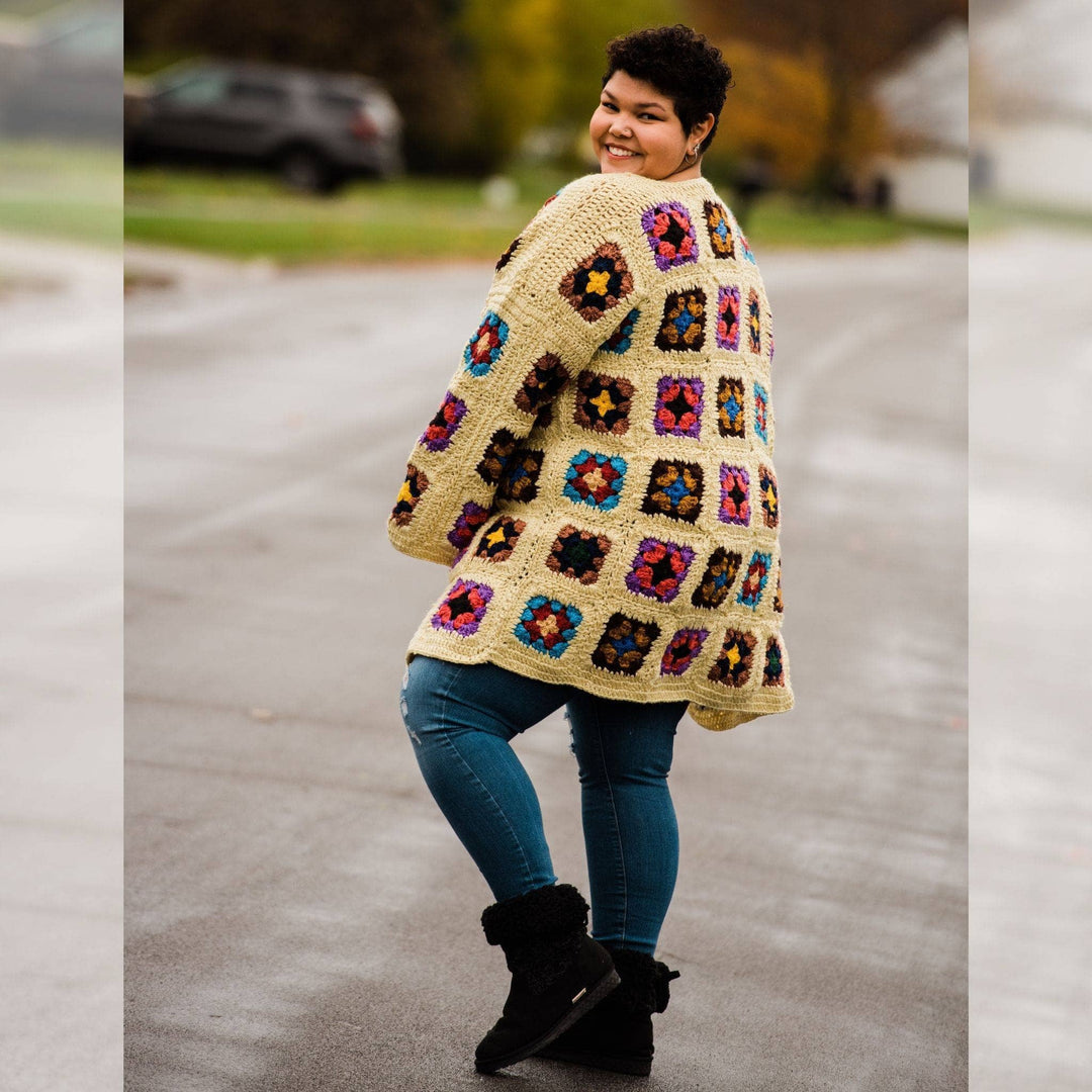 Back view of model wearing goddess size technicolor long cardigan (crochet granny square, beige and multicolor) while standing outside on a quiet street.