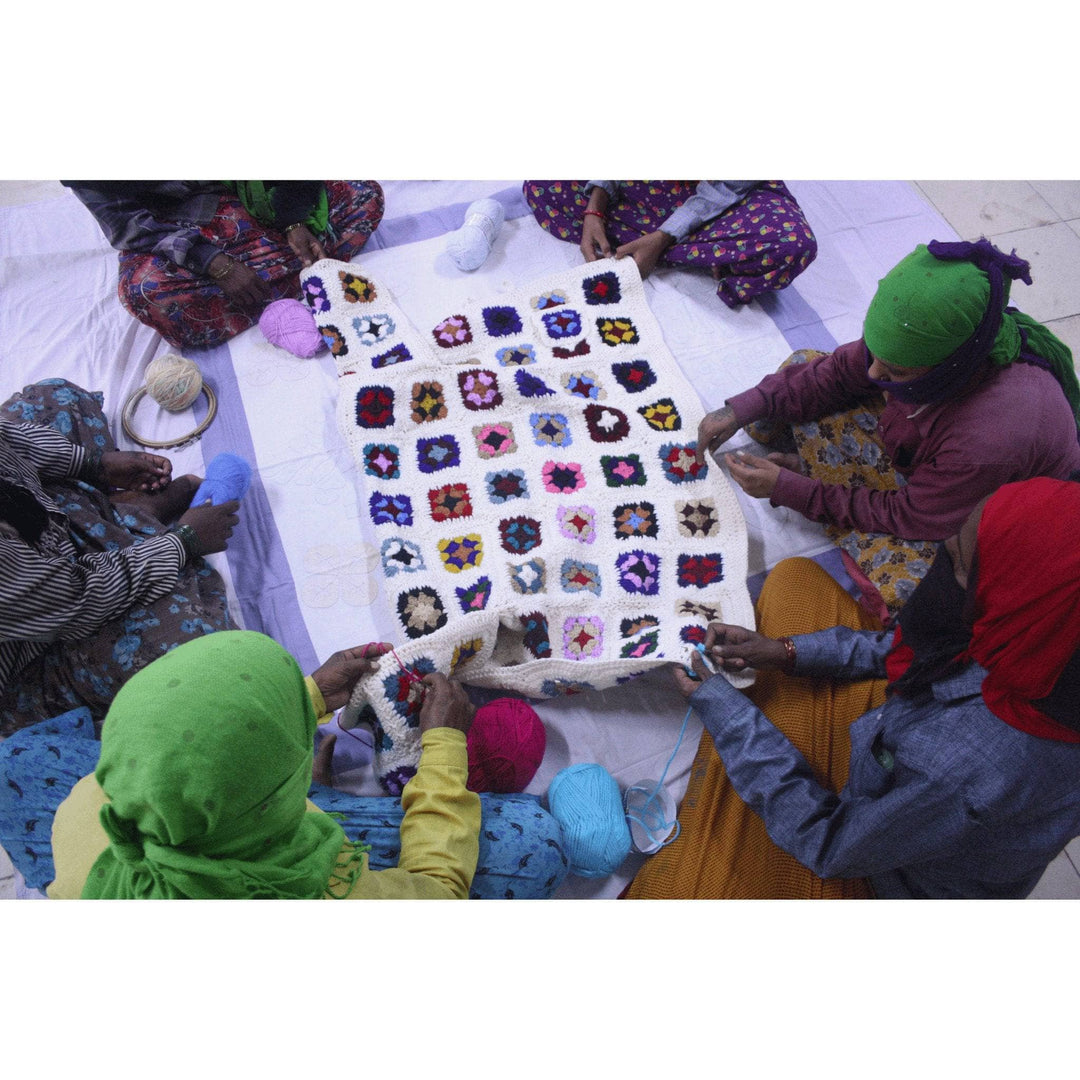 6 women sitting in a circle around a panel of a granny square long cardigan working on it together.