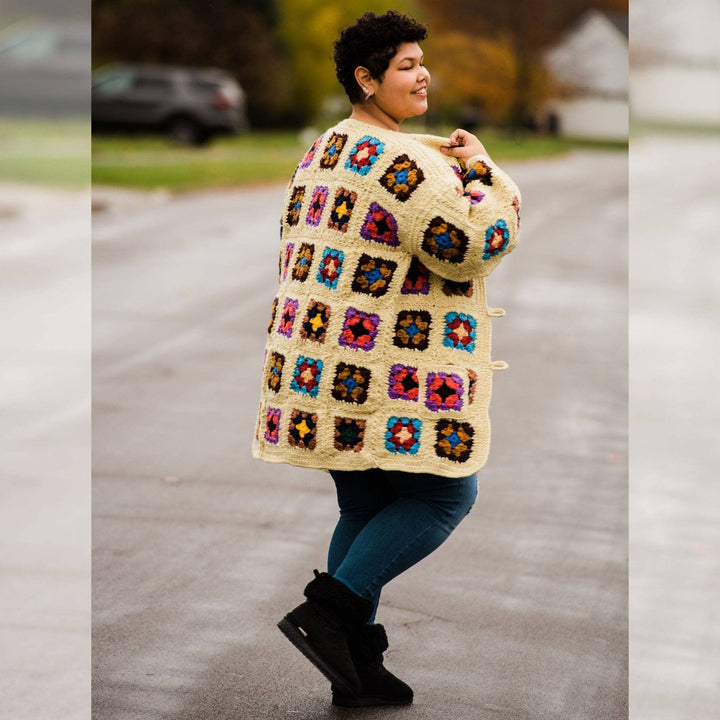 Back view of Model wearing goddess size technicolor long cardigan (crochet granny square, beige and multicolor) while standing outside on a quiet street.