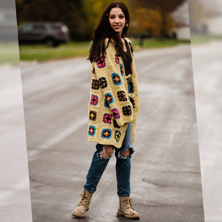 Model wearing regular size technicolor long cardigan (crochet granny square, beige and multicolor) while standing outside on a quiet street.