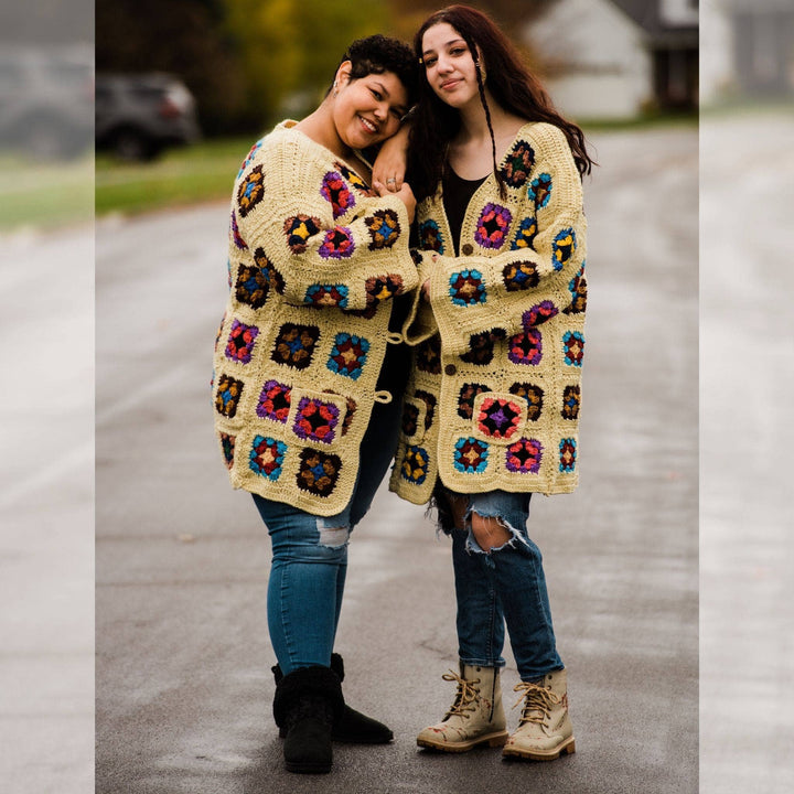 Models wearing technicolor long cardigan (crochet granny square, beige and multicolor) Left goddess and right regular while standing outside on a quiet street.