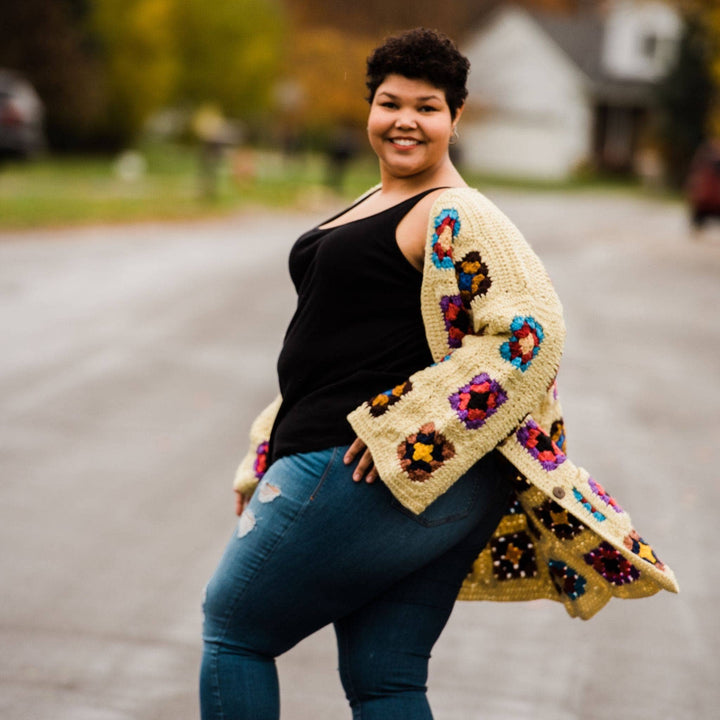 Model wearing goddess size technicolor long cardigan (crochet granny square, beige and multicolor) while standing outside on a quiet street.