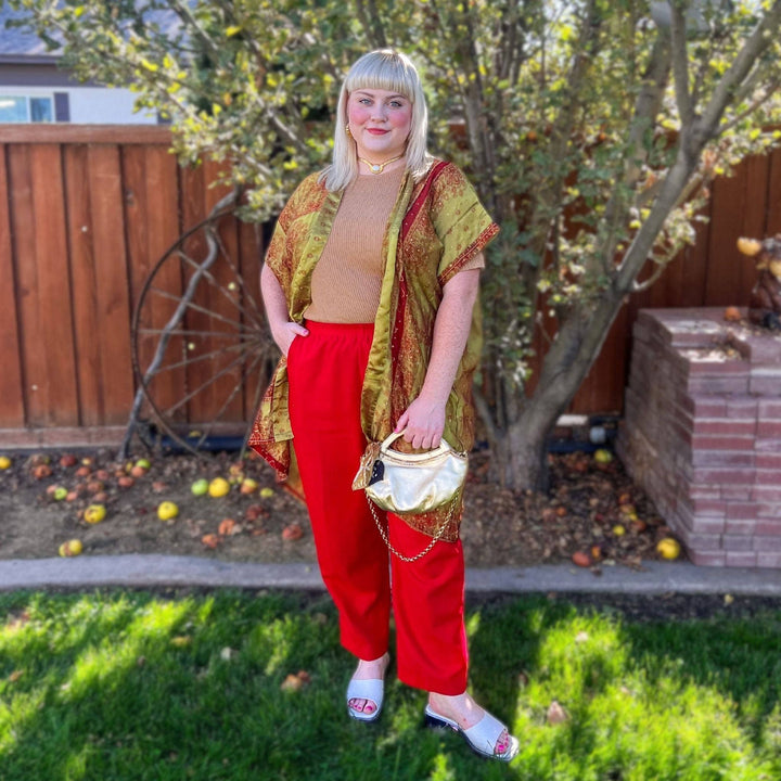 A model wearing a short amara sari duster in her backyard. The Duster is gold with red trim and accents. She's pair the duster with a cream colored top and red pants.