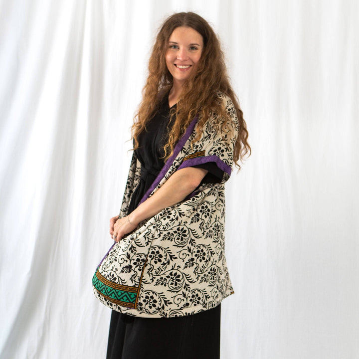 A woman standing in front of a white background with a white sari silk duster over a black dress. The Duster has delicate black floral patterns all over with purple edging.