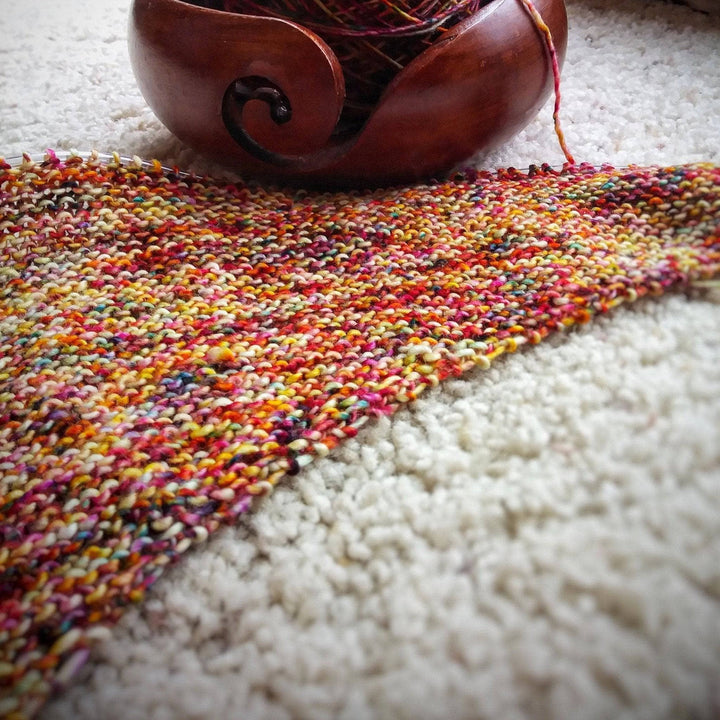 In progress picture of a red and yellow speckled shawl with a yarn bowl in the top of the picture on a white background