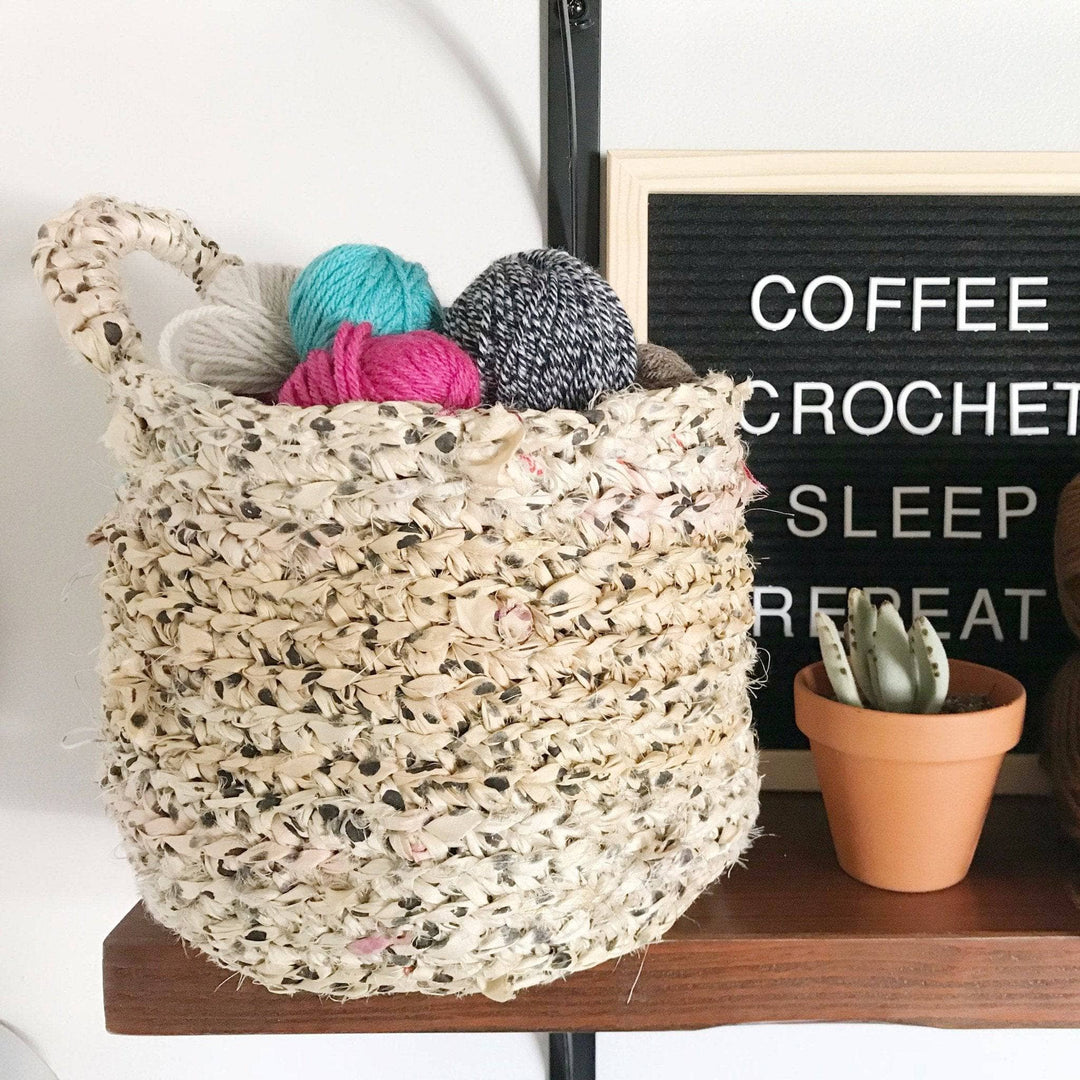 The Hayden Hanging Basket holding balls of yarn and sitting on a wooden shelf next to a potted plant and black letterboard