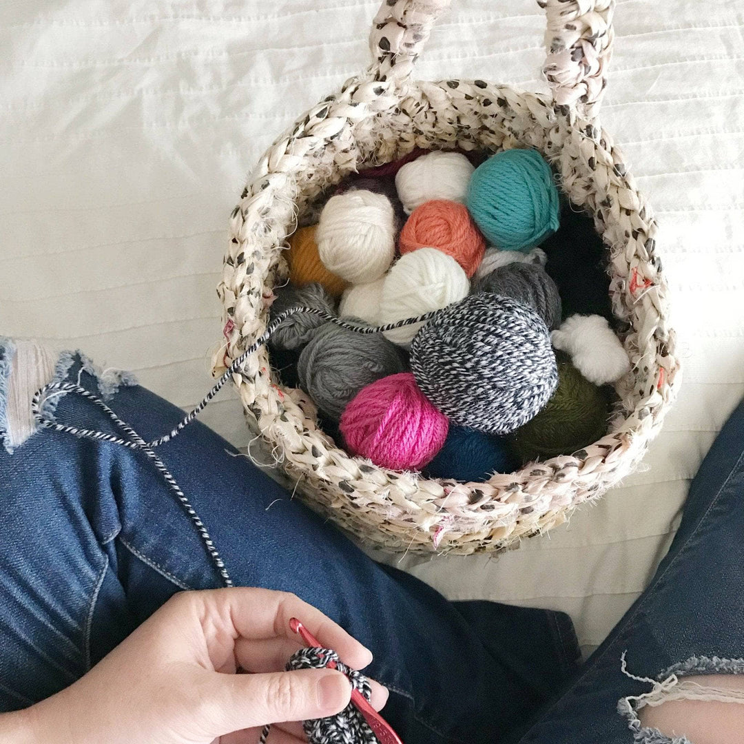The Hayden Hanging Basket holding balls of yarn and sitting a white bed with woman's hands working on a gray crochet project