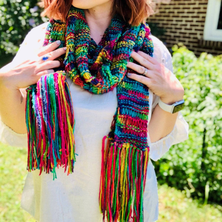 model wering knit rainbow scarf with greenery in the background.