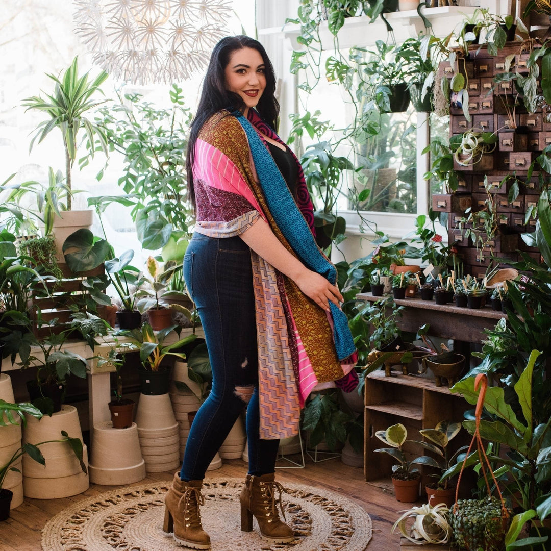 Model is wearing a pink and blue medley scarf around their shoulders in front of potted plants.  