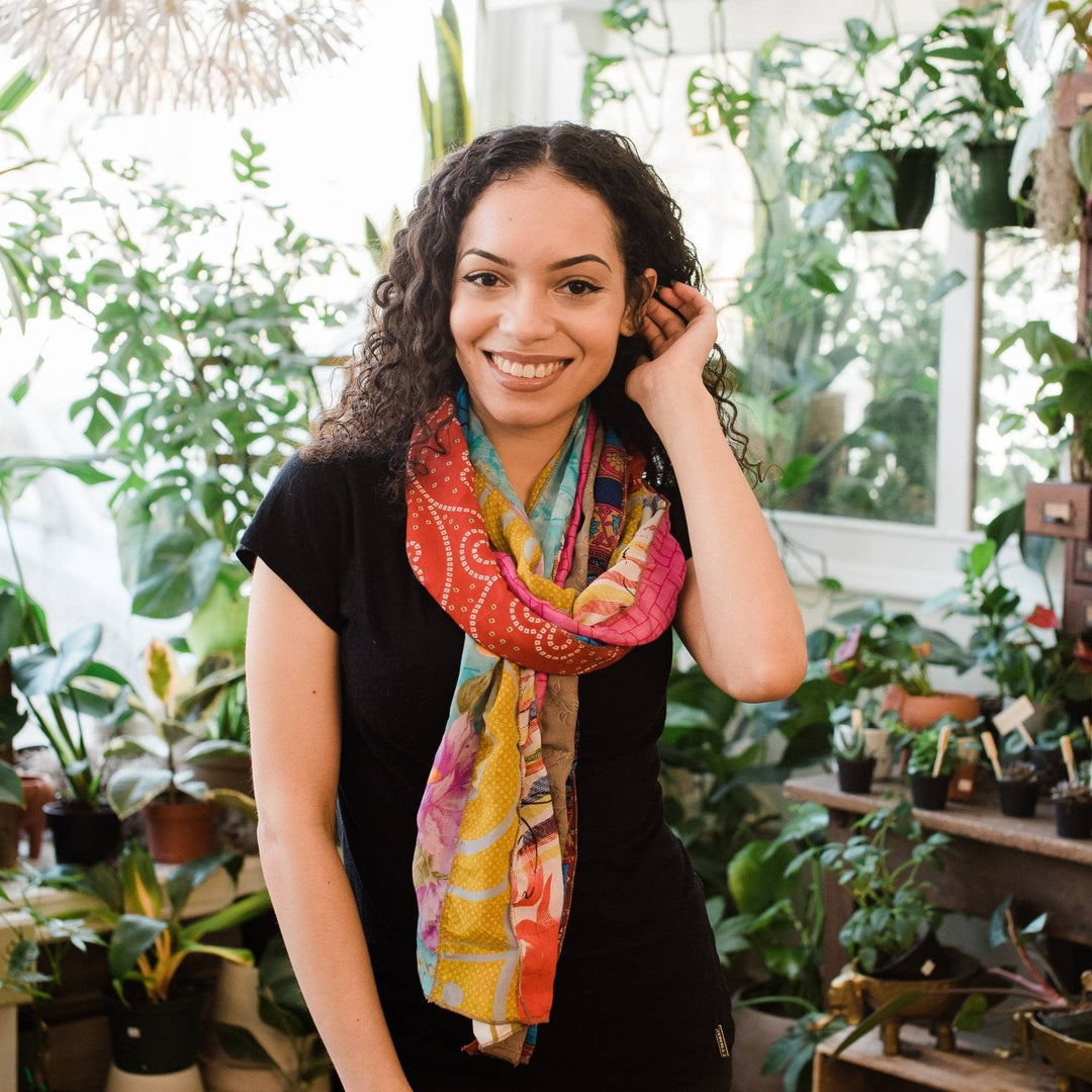 Model is wearing a brightly colored sari silk scarf around their neck with potted plants in the background. 