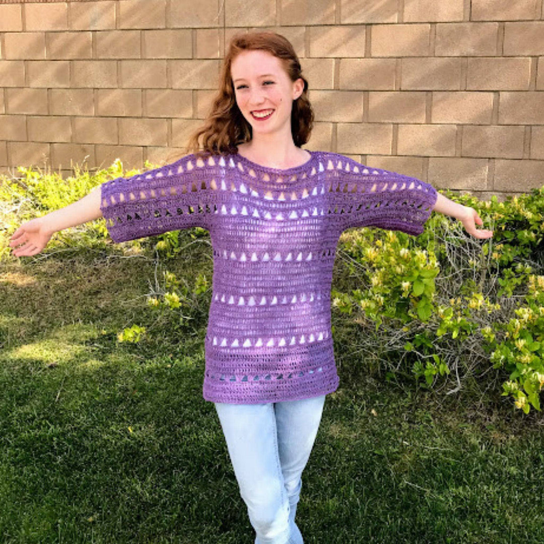 A woman standing in front of a bush with her arms out. She's wearing a shelby crochet top made using purple 2-ply linen yarn.